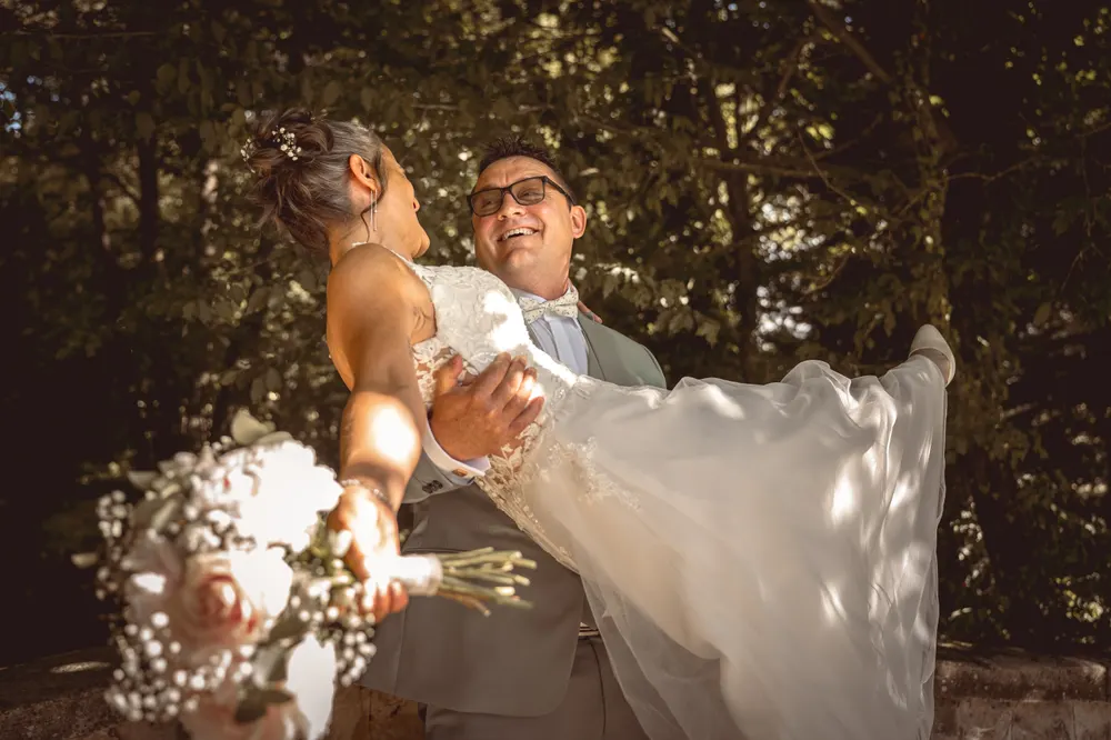 Photo de mariage naturelle à Salins (77) : le marié porte sa mariée dans ses bras en riant, sous les arbres