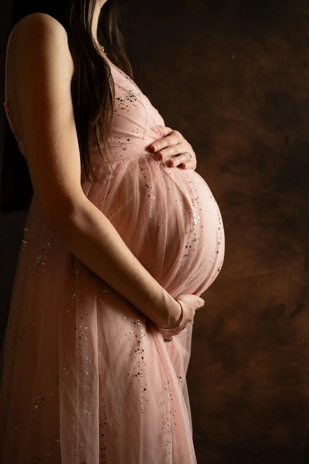 Photographe grossesse à Salins (77) : femme enceinte en robe rose pailletée tenant son ventre, sur fond sombre au studio Maison Rischard