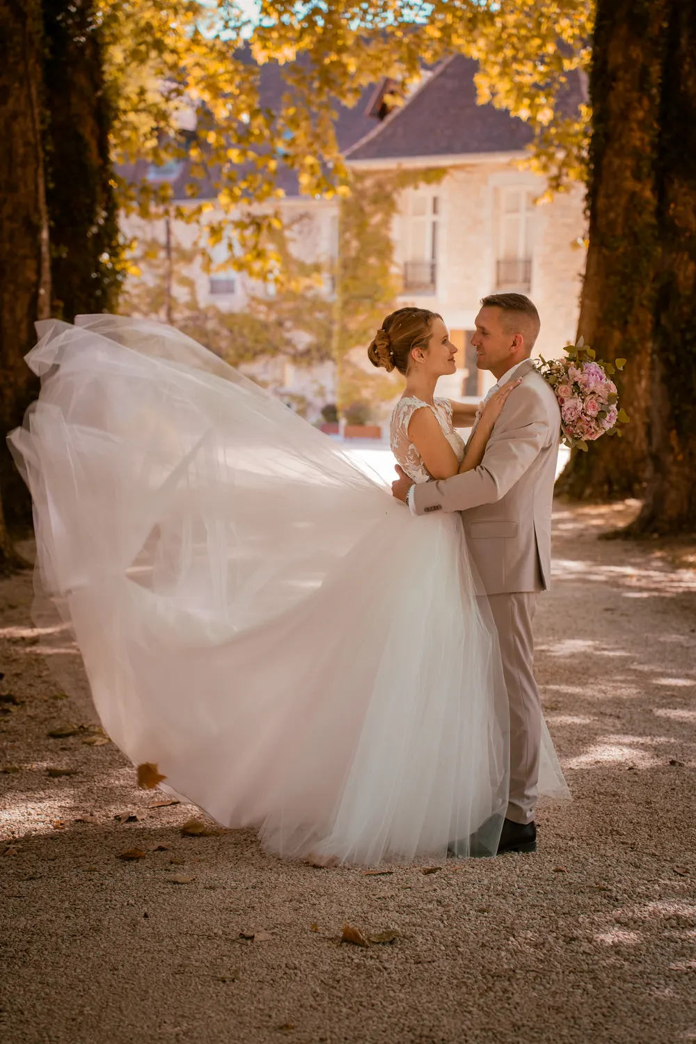 Photographe mariage en Seine-et-Marne : les mariés enlacés dans une allée boisée, le voile de la mariée soulevé par le vent