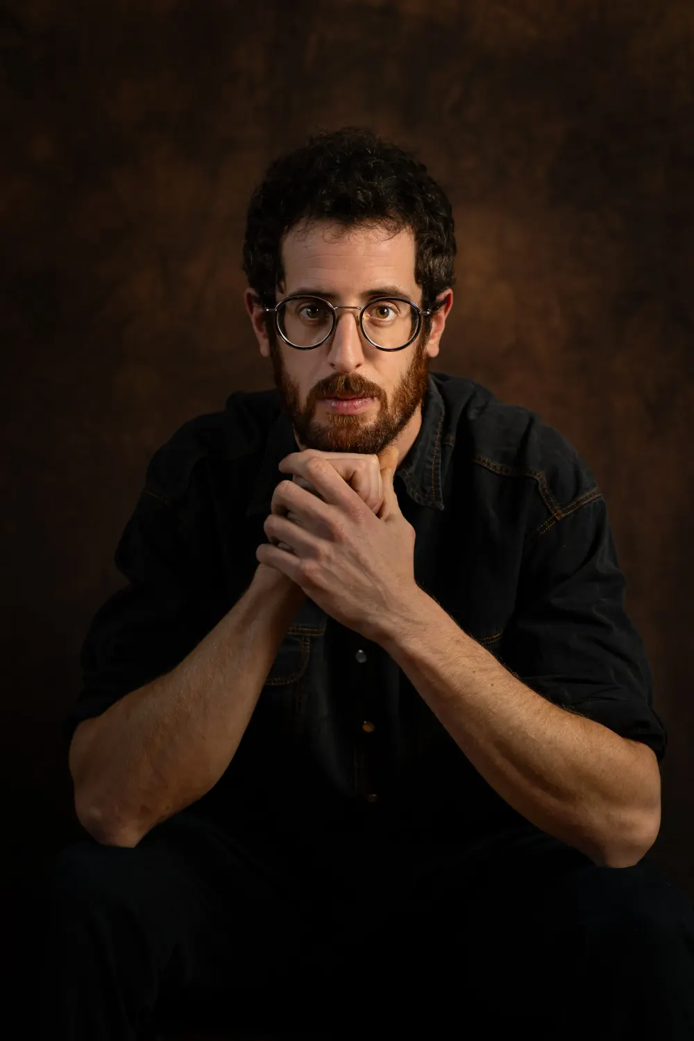 Séance photo portrait artistique en Seine-et-Marne : homme barbu à lunettes rondes, mains sous le menton, sur fond sombre