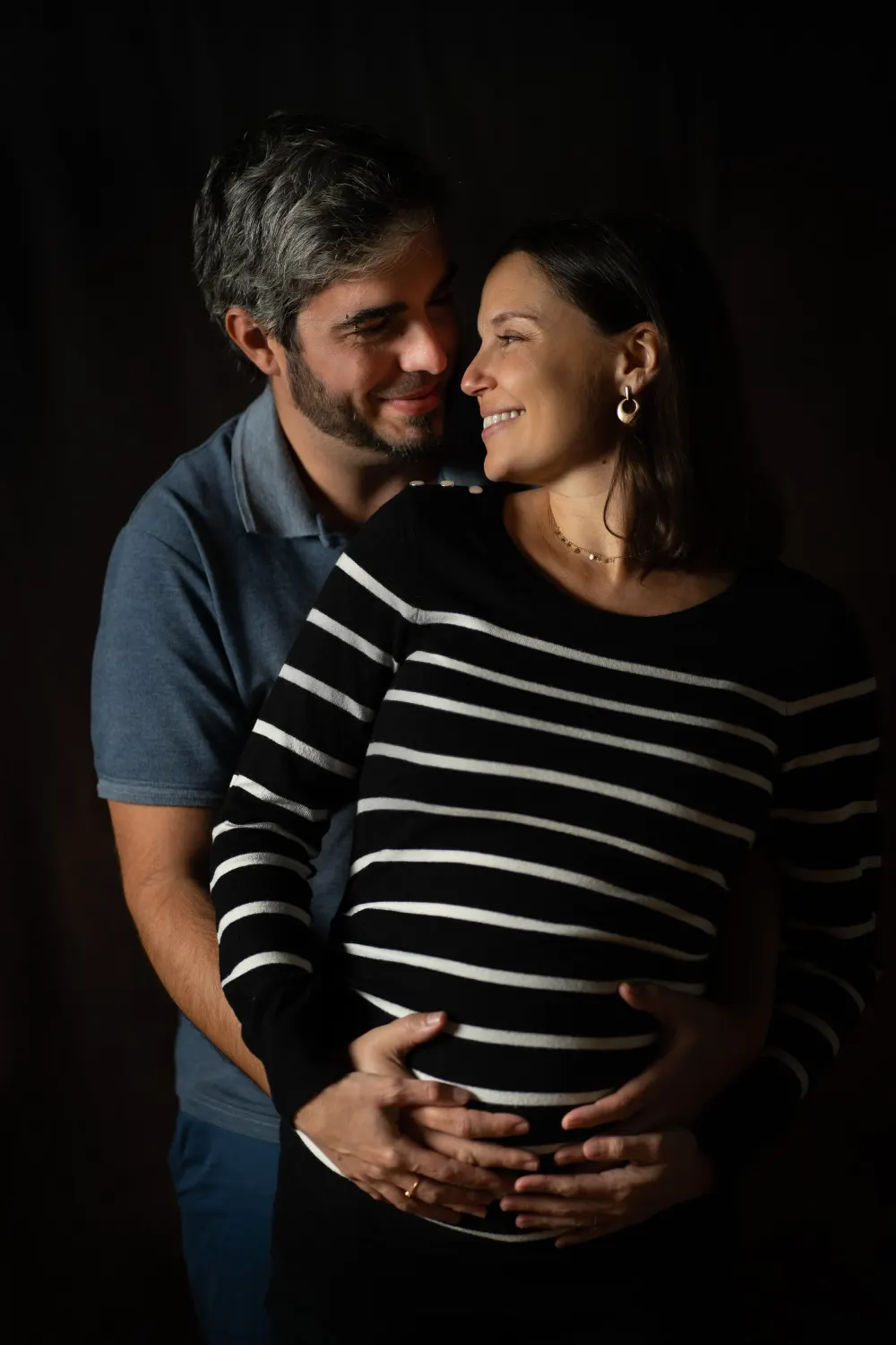 Photographe maternité à Salins (77) : un couple pose souriant derrière une femme enceinte en pull rayé
